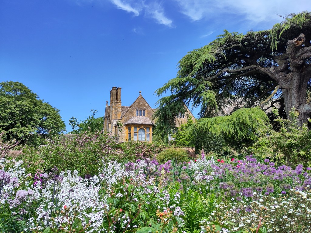 An arts and crafts style manor house in honey coloured Cotswold stone sits above a full garden, the predominant colours are white,pinks and purples. 