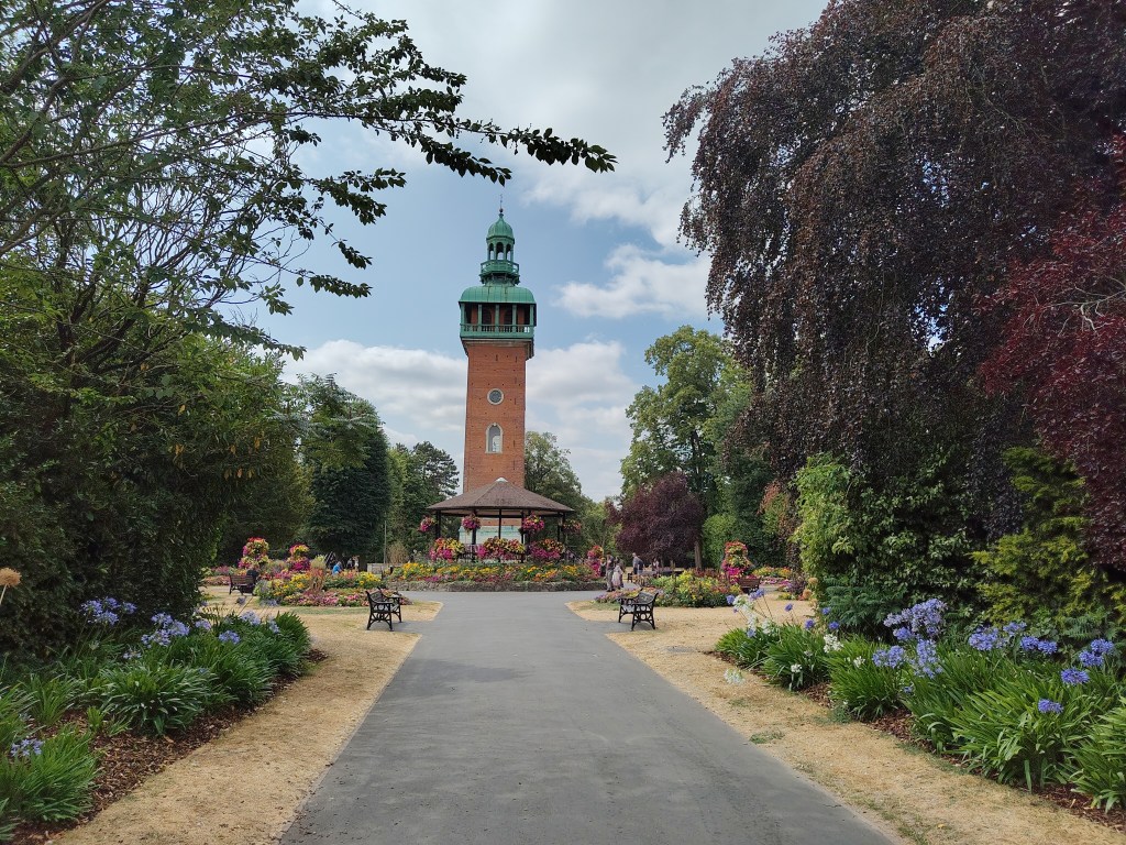 A red brick tower with copper top storey. At the bottom is a Victorian style bandstand with hanging baskets planted up with colourful flowers.