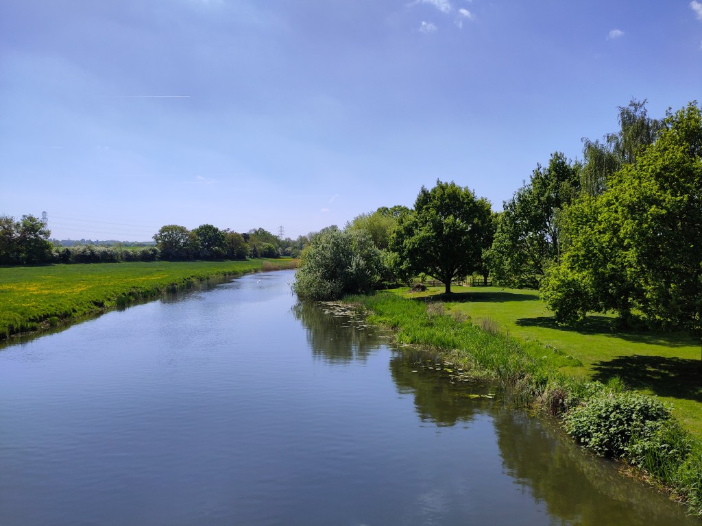 A wide calm river lined on both sides by green fields and trees.
